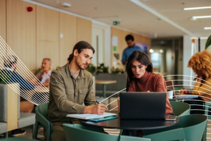 Two people are seated at a round table in a modern indoor setting, working together on a laptop. One person is writing in a notebook while the other is focused on the laptop screen.