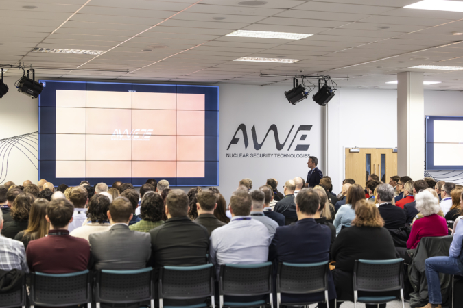 Group of people sat down facing away from the camera sat in rows at a conference watching a stage with a large screen