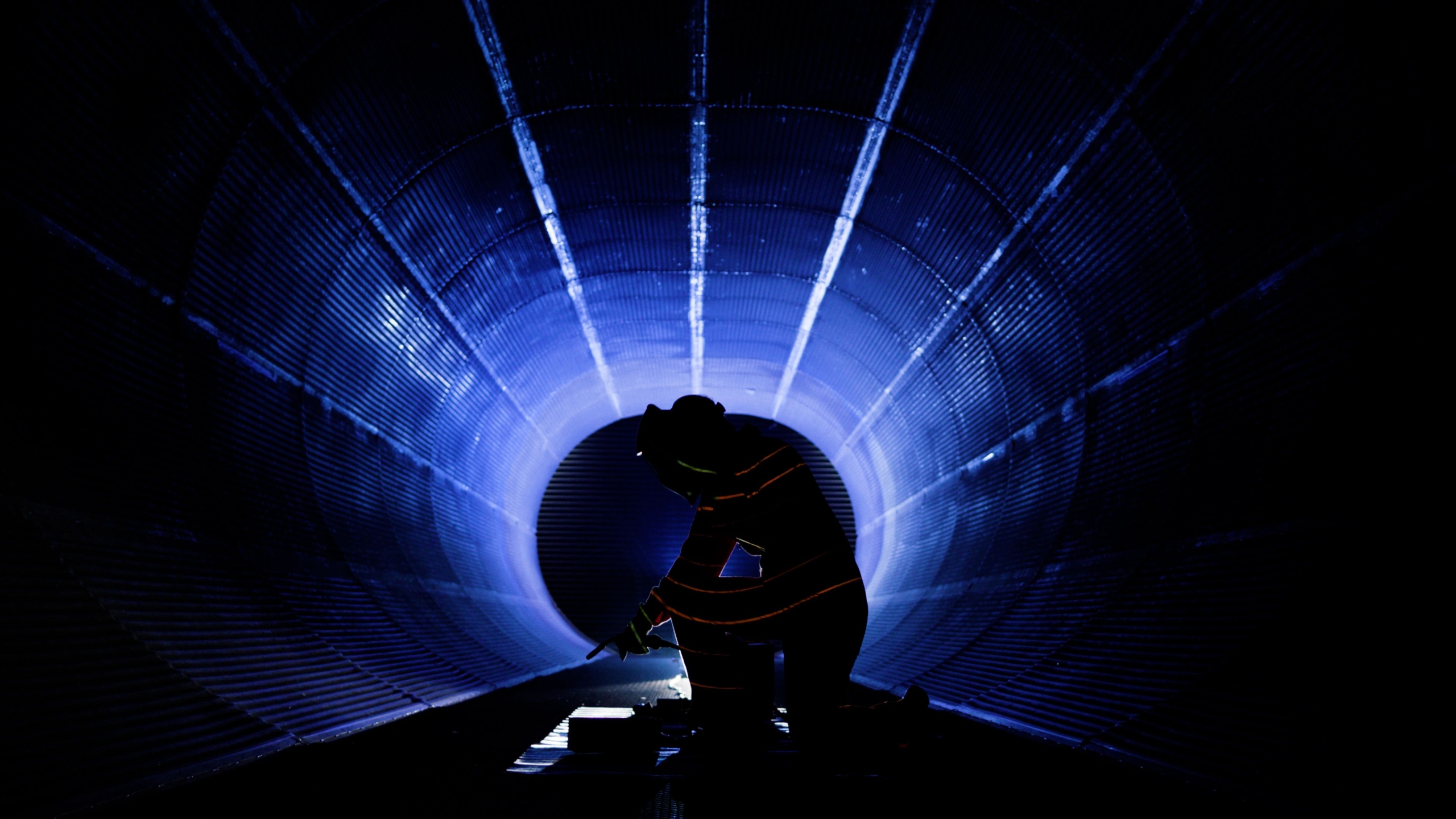 Silhouette of a person working inside a large cylindrical tunnel, illuminated by blue light beams converging toward the center. The figure is kneeling and engaged in a technical task, creating a dramatic industrial scene.