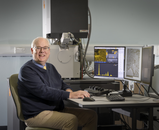 Professor Mark Whiting seated at a desk operates scientific equipment, with two computer monitors displaying microscopy images and analysis software. A large laboratory instrument is positioned behind the desk, and various control devices, including a keyboard and specialized control panel, are placed in front of the person.